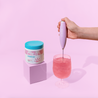 Cira Nutrition Pink Frother with Cira logo. A woman's hand is stirring Watermelon Sugar Hydrate next to a tub of Cira Nutrition Watermelon Sugar Hydrate in a white tub with a pink label and blue lid. The background is pink. 